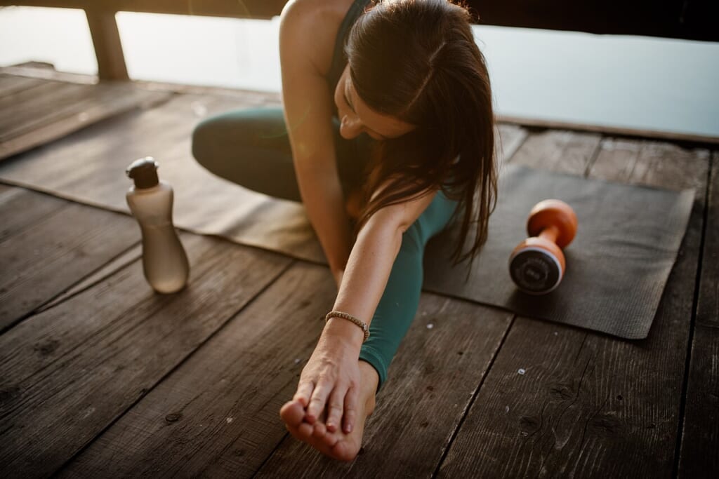 Woman stretching on yoga mat near dumbbell and water bottle | オンザショア立川店 立川駅徒歩1分年中無休の溶岩ホットヨガスタジオ 木の床に敷いたヨガマットで体を伸ばす女性とマットの上のダンベルと水筒 立川駅徒歩1分年中無休の溶岩ホットヨガスタジオ