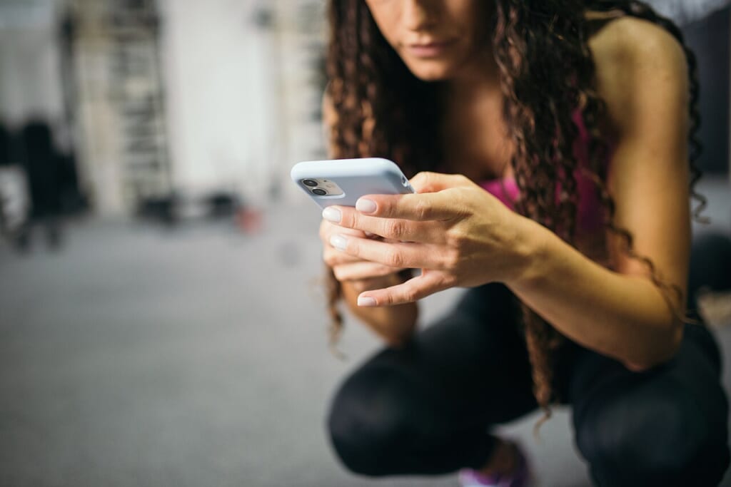 Young woman using a smartphone in the gym | オンザショア立川店 立川駅徒歩1分年中無休の溶岩ホットヨガスタジオ ジムの中でスマートフォンを手に持ち使用している女性の様子を写した写真 立川駅徒歩1分年中無休の溶岩ホットヨガスタジオ