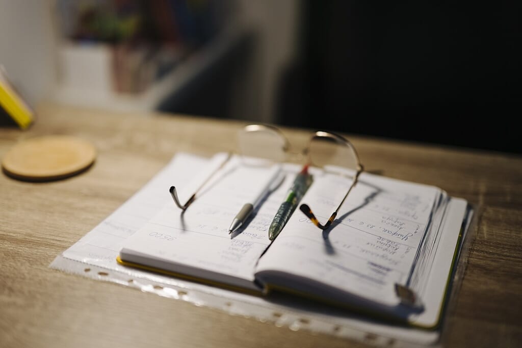 Desk with notebook pens and glasses | オンザショア立川店 立川駅徒歩1分年中無休の溶岩ホットヨガスタジオ 机の上に開かれたノートと数本のペンが置かれノートの上には眼鏡が載っている 立川駅徒歩1分年中無休の溶岩ホットヨガスタジオ