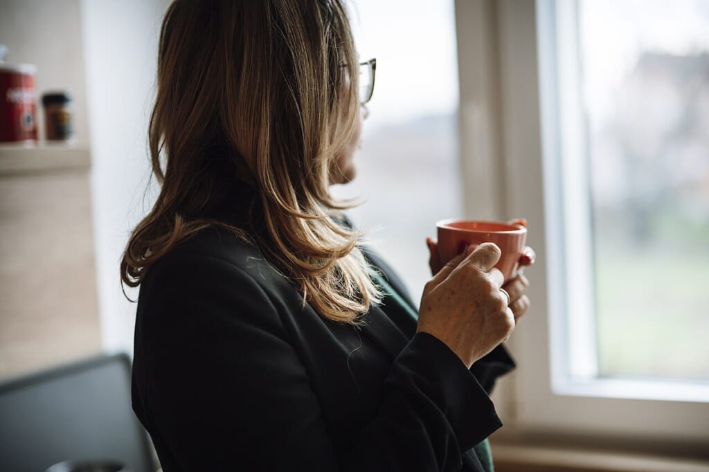 Woman enjoys hot drink while looking out window in kitchen during daytime at home | オンザショア立川店 立川駅徒歩1分年中無休の溶岩ホットヨガスタジオ 窓から光が入るキッチンでカップを両手で持ちながら窓の外を見る女性 立川駅徒歩1分年中無休の溶岩ホットヨガスタジオ