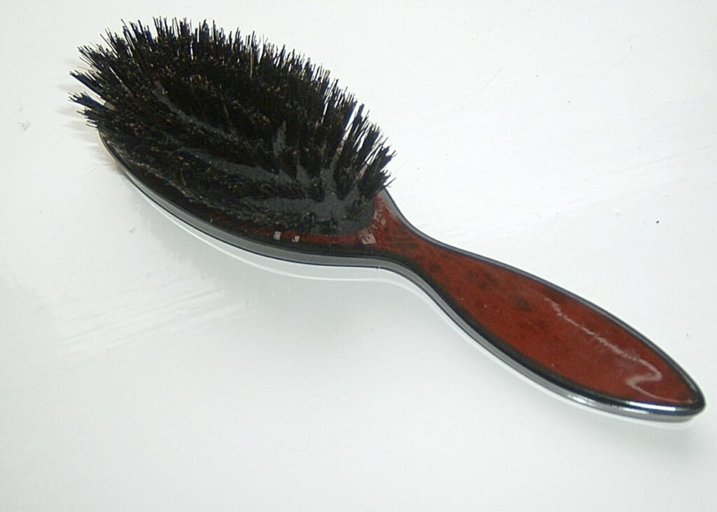 白い台の上に置かれた黒い毛を密に植えた赤茶色の柄のヘアブラシ1本 立川駅徒歩1分年中無休の溶岩ホットヨガスタジオ
