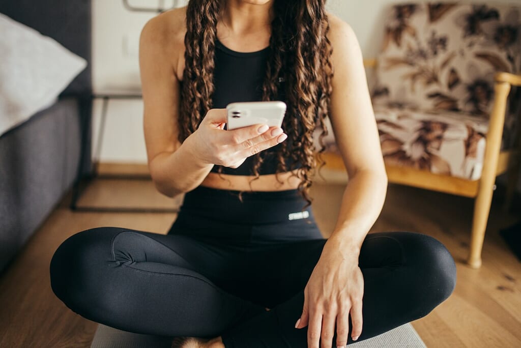 Woman sitting on a yoga mat with her legs crossed and checking her phone | オンザショア立川店 立川駅徒歩1分年中無休の溶岩ホットヨガスタジオ 黒いレギンス姿でヨガマットにあぐらをかき手に持ったスマートフォンを見る女性 立川駅徒歩1分年中無休の溶岩ホットヨガスタジオ