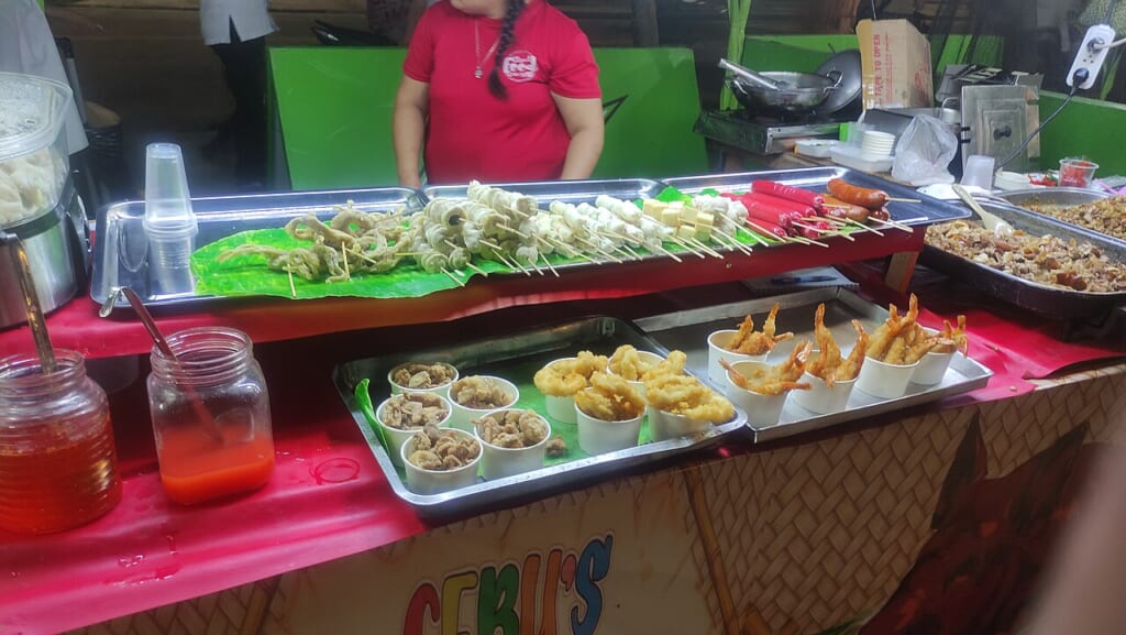 Fried Street Snacks Displayed | オンザショア立川店 立川駅徒歩1分年中無休の溶岩ホットヨガスタジオ 金属製のトレーに並ぶ串刺しの軽食と紙カップに盛られたイカの揚げ物がある屋台 立川駅徒歩1分年中無休の溶岩ホットヨガスタジオ