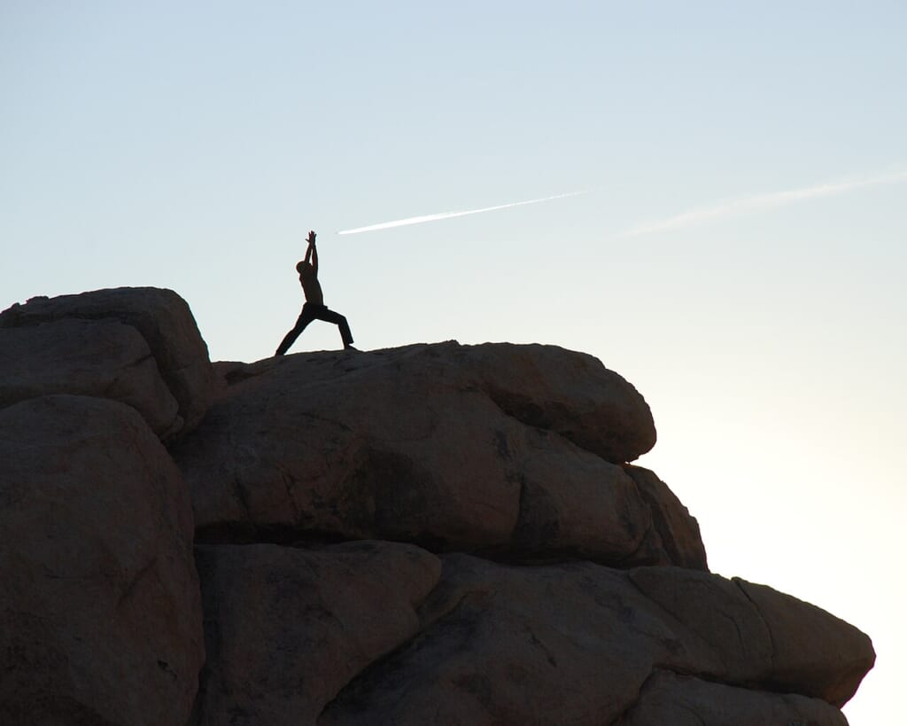 Joshua Tree yoga warrior 1a | オンザショア立川店 立川駅徒歩1分年中無休の溶岩ホットヨガスタジオ 朝日の屋外で立った姿勢でヨガのウォーリアーⅠのポーズをとる人 立川駅徒歩1分年中無休の溶岩ホットヨガスタジオ