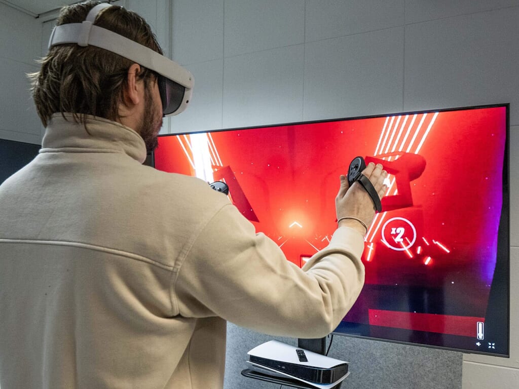 InclusiveGameLab PersonPlayingVirtualReality 11 CCBYSA | オンザショア立川店 立川駅徒歩1分年中無休の溶岩ホットヨガスタジオ 大きな画面の前に立ちVRヘッドセットを装着して右手にコントローラーを持つ人 立川駅徒歩1分年中無休の溶岩ホットヨガスタジオ