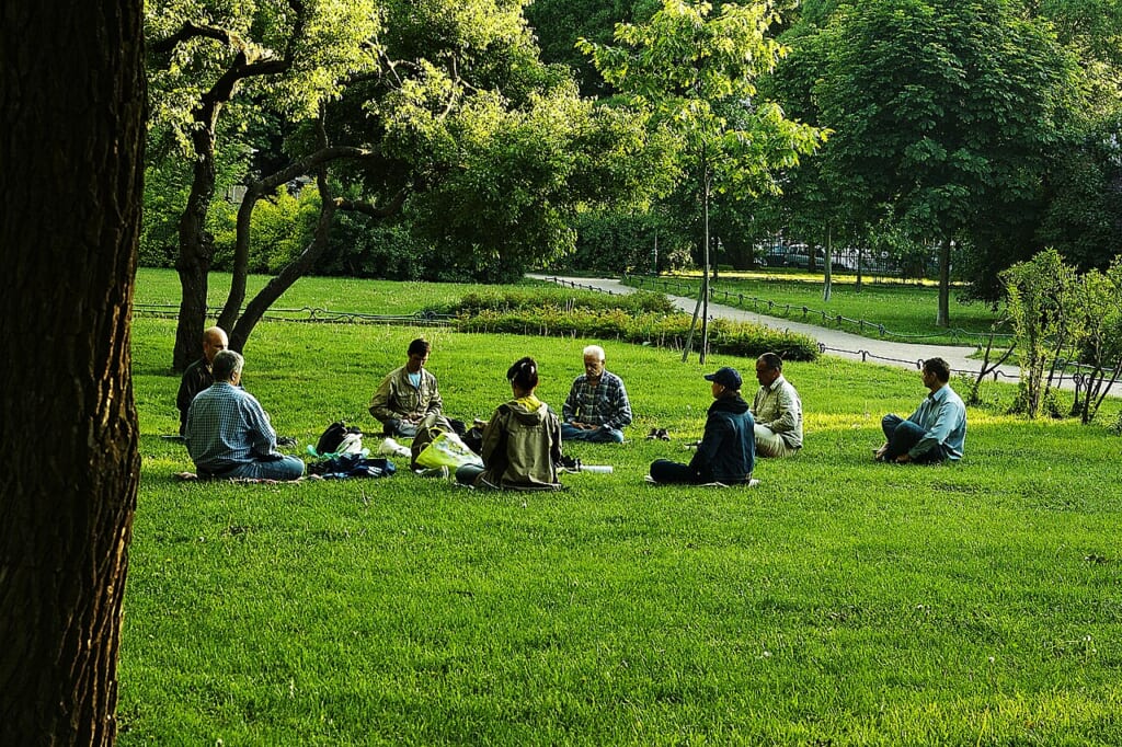 A group meditation session in a park in a yoga asana | オンザショア立川店 立川駅徒歩1分年中無休の溶岩ホットヨガスタジオ 公園で集まり座位のヨガの姿勢をとって瞑想に取り組む人たちの様子 立川駅徒歩1分年中無休の溶岩ホットヨガスタジオ
