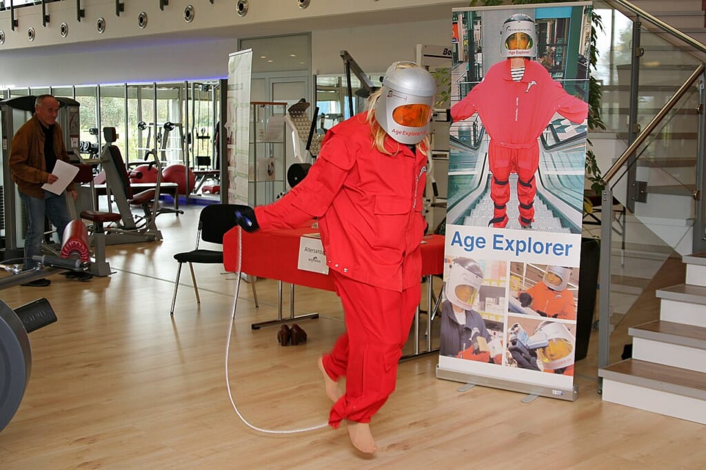 Skipping rope agesuit altersanzug alterssimulationsanzug ageexplorer gundolf meyerhentschel | オンザショア立川店 立川駅徒歩1分年中無休の溶岩ホットヨガスタジオ 赤い加齢体験スーツを着た女性が縄を使って縄跳びを試している様子 立川駅徒歩1分年中無休の溶岩ホットヨガスタジオ
