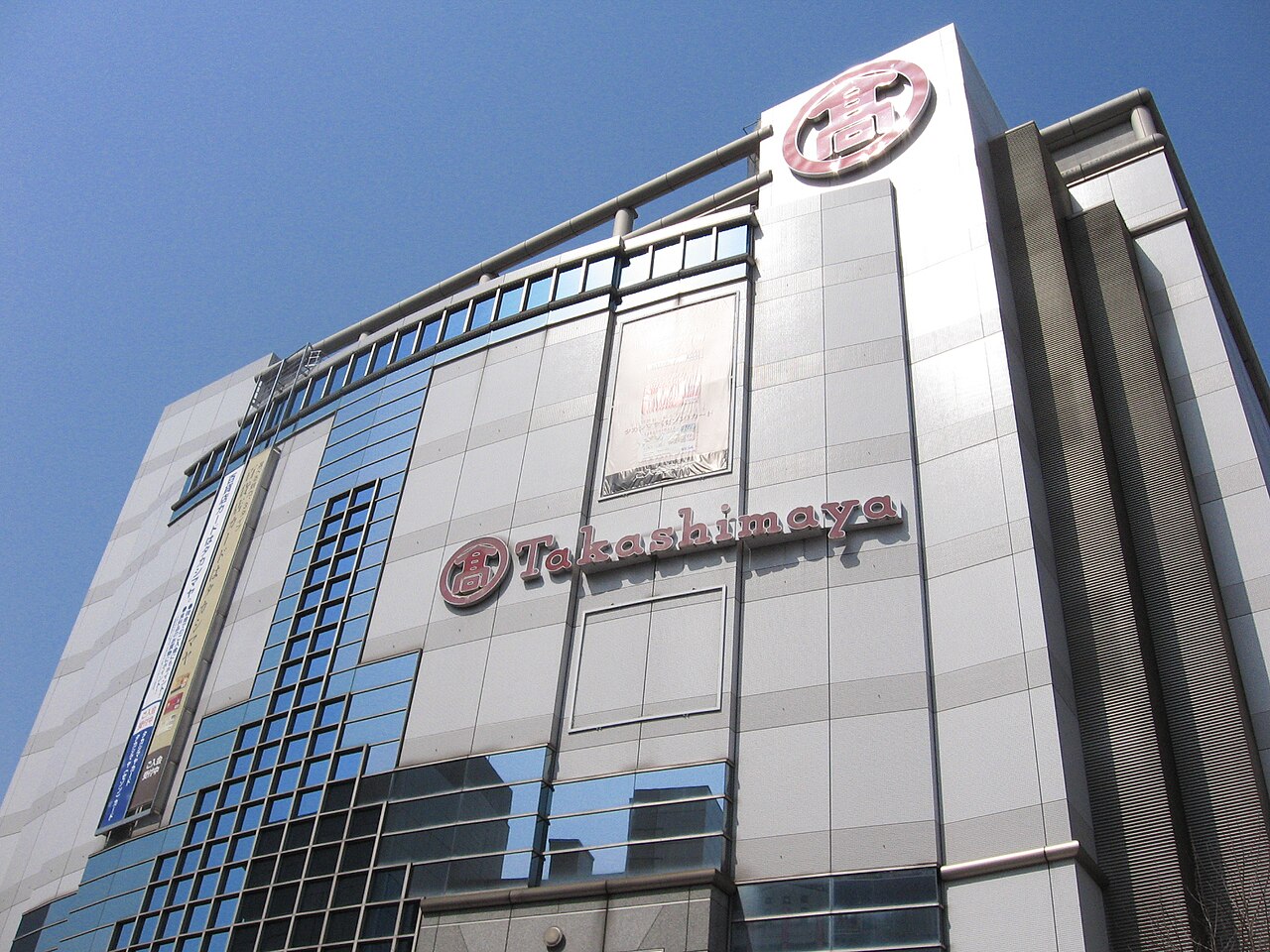 Looking up at the Tachikawa Takashimaya building, with the Takashimaya sign and crest on its wall.