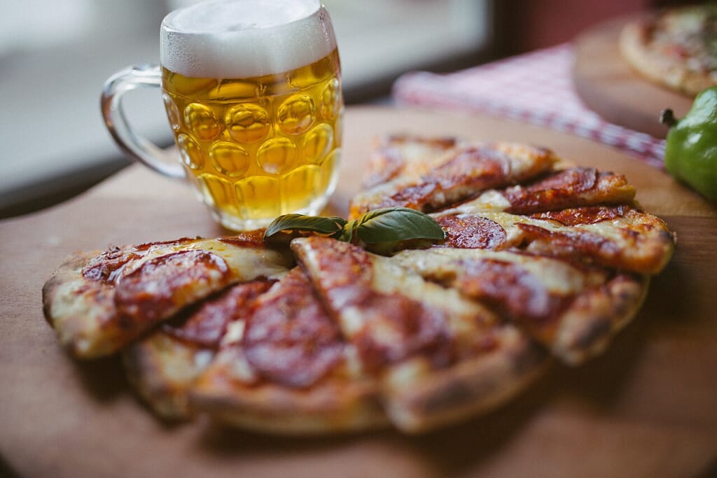 Beer and pizza on display in a restaurant on a rustic table with focus on the glass of beer | オンザショア立川店 立川駅徒歩1分年中無休の溶岩ホットヨガスタジオ 木のテーブルに置かれたビールのグラスとピザビールのグラスに焦点を合わせた写真 立川駅徒歩1分年中無休の溶岩ホットヨガスタジオ