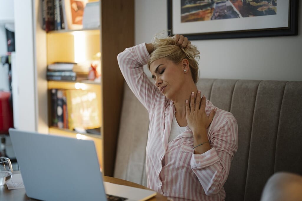 Stressed woman in home office takes break to stretch and relieve neck tension during work hours | オンザショア立川店 立川駅徒歩1分年中無休の溶岩ホットヨガスタジオ ノートパソコンのある机の前で椅子に座りながら首を伸ばしている女性 立川駅徒歩1分年中無休の溶岩ホットヨガスタジオ