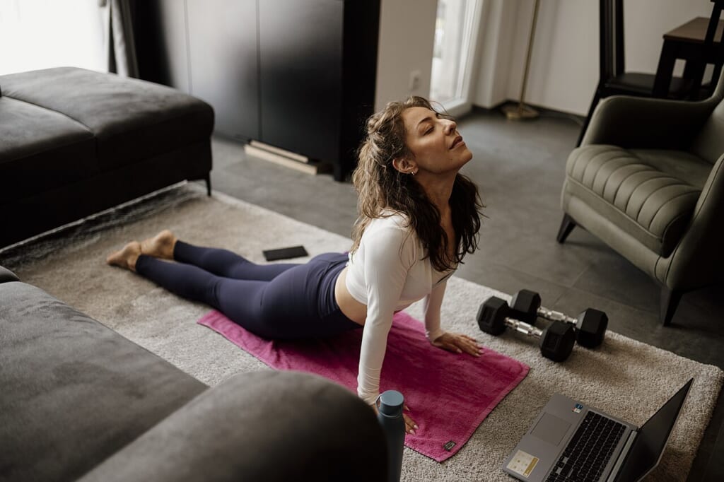 Woman practicing yoga at home on a mat in a living room | オンザショア立川店 立川駅徒歩1分年中無休の溶岩ホットヨガスタジオ 明るい居間のマットの上でそばにノートパソコンを置き頭を後ろに傾けてヨガの姿勢をとる女性 立川駅徒歩1分年中無休の溶岩ホットヨガスタジオ