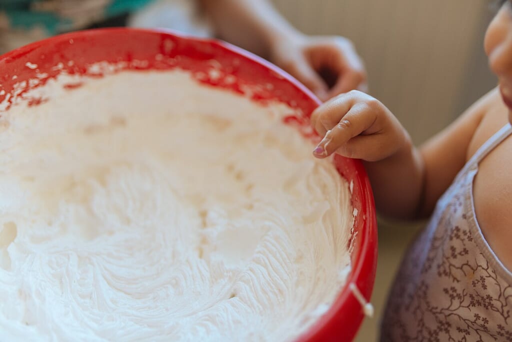 Childs hand reaching into a bowl of whipped cream in a kitchen during a family cooking time | オンザショア立川店 立川駅徒歩1分年中無休の溶岩ホットヨガスタジオ 台所でホイップクリームの入った大きな赤いボウルに手を伸ばす子どもの手元 立川駅徒歩1分年中無休の溶岩ホットヨガスタジオ