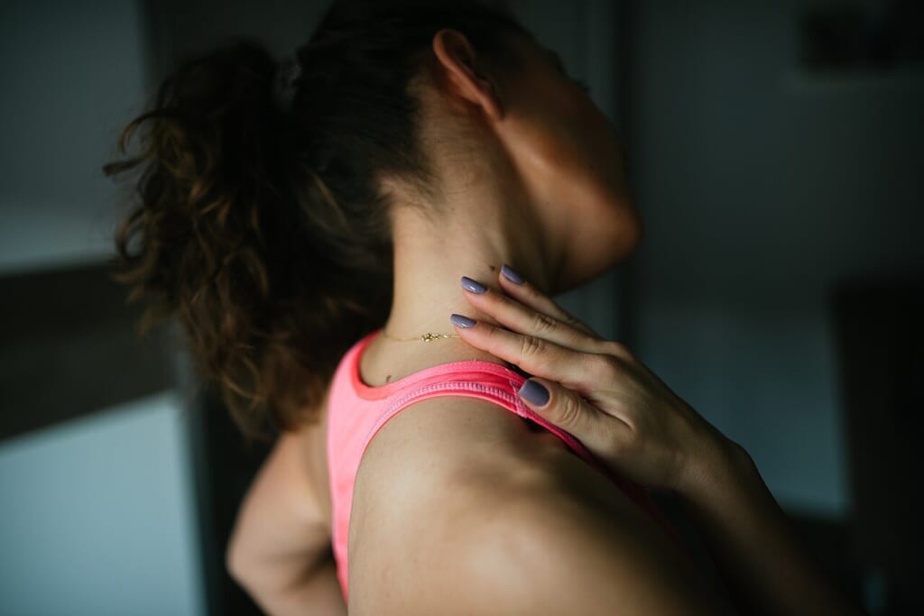 A woman in a pink athletic top is gently stretching her neck indoors with her back partially turned | オンザショア立川店 立川駅徒歩1分年中無休の溶岩ホットヨガスタジオ ピンクの運動着を着た女性が室内で背中を斜めに向けて首をストレッチしている 立川駅徒歩1分年中無休の溶岩ホットヨガスタジオ