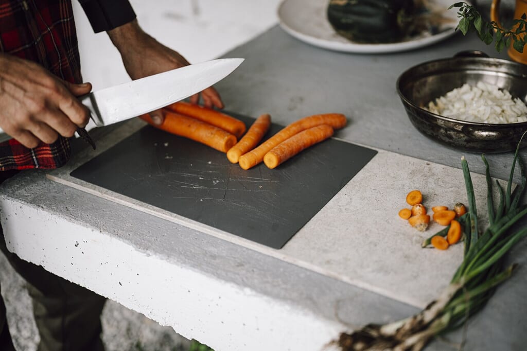 Preparing fresh carrots for a healthy meal in a rustic kitchen setting | オンザショア立川店 立川駅徒歩1分年中無休の溶岩ホットヨガスタジオ 台所の石のまな板でにんじんを切る手元とそのそばに並べられた野菜 立川駅徒歩1分年中無休の溶岩ホットヨガスタジオ