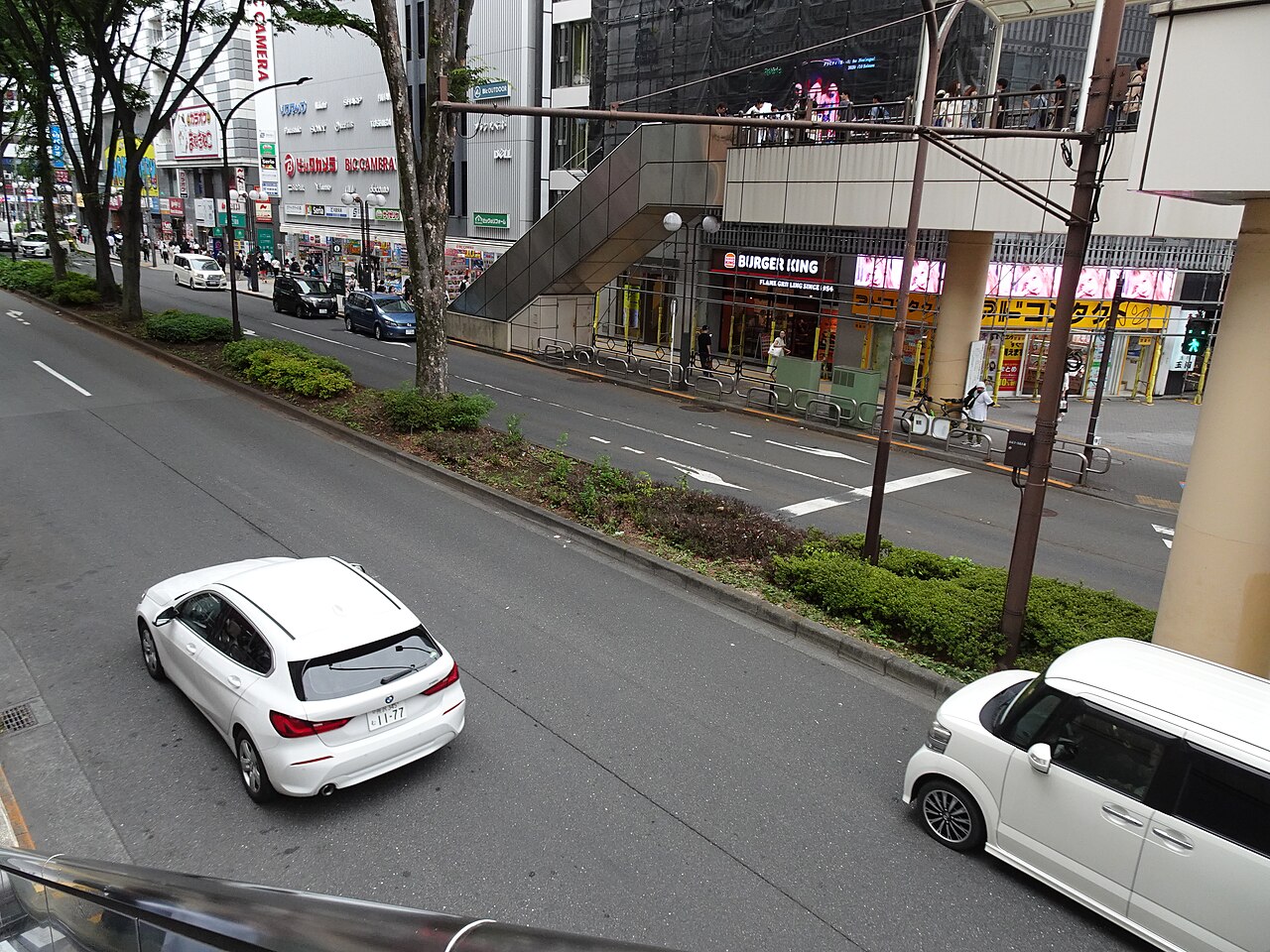 North Exit Avenue seen from the pedestrian deck north of Tachikawa Station, cars passing under the trees (2026)