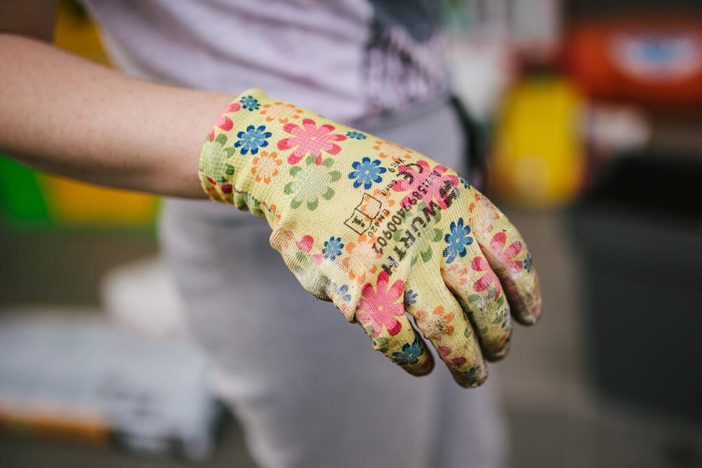 A hand wearing bright floral gardening gloves | オンザショア立川店 立川駅徒歩1分年中無休の溶岩ホットヨガスタジオ 花柄の園芸用手袋を着けた手で共同菜園に花を植える準備をしている様子 立川駅徒歩1分年中無休の溶岩ホットヨガスタジオ