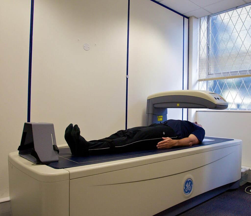 DEXA scanner in use ALSPAC | オンザショア立川店 立川駅徒歩1分年中無休の溶岩ホットヨガスタジオ 検査台に横たわる男性の体の上にDXA検査装置のアームが配置されている様子 立川駅徒歩1分年中無休の溶岩ホットヨガスタジオ