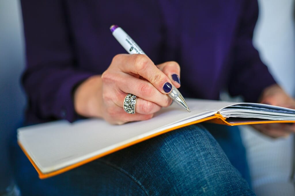 Woman writing on a notebook with a pen | オンザショア立川店 立川駅徒歩1分年中無休の溶岩ホットヨガスタジオ ノートを開きペンを使ってページに文字を書き込んでいる女性の様子 立川駅徒歩1分年中無休の溶岩ホットヨガスタジオ