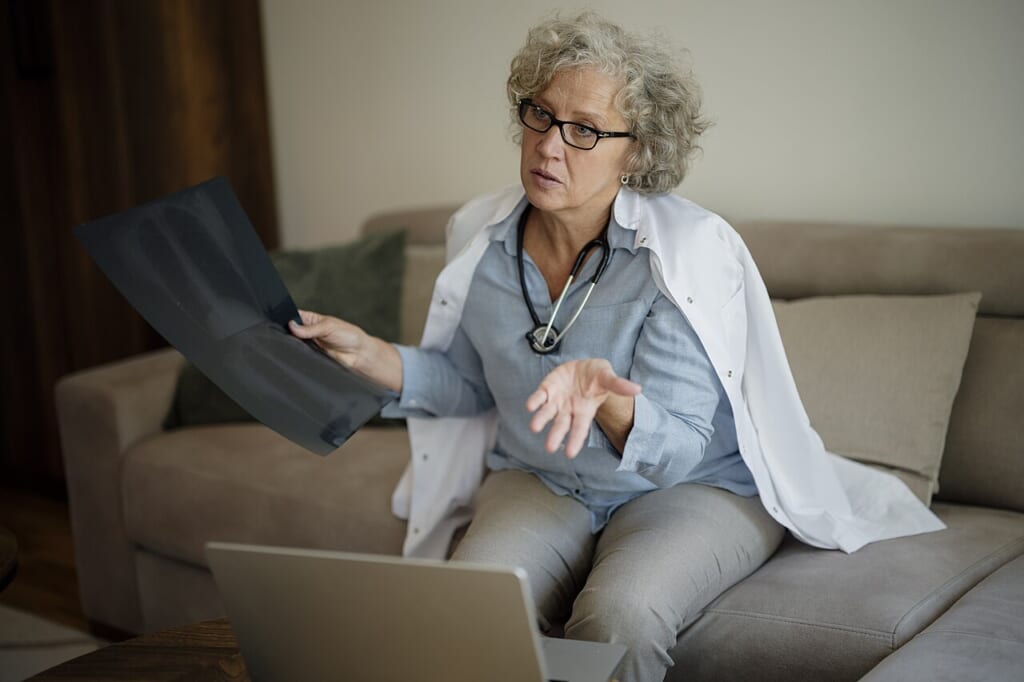 Doctor discussing medical images at home in a cozy living room during a virtual consultation | オンザショア立川店 立川駅徒歩1分年中無休の溶岩ホットヨガスタジオ ソファに座り手に持った医療画像を示しながらノートパソコンを前に話す医師 立川駅徒歩1分年中無休の溶岩ホットヨガスタジオ