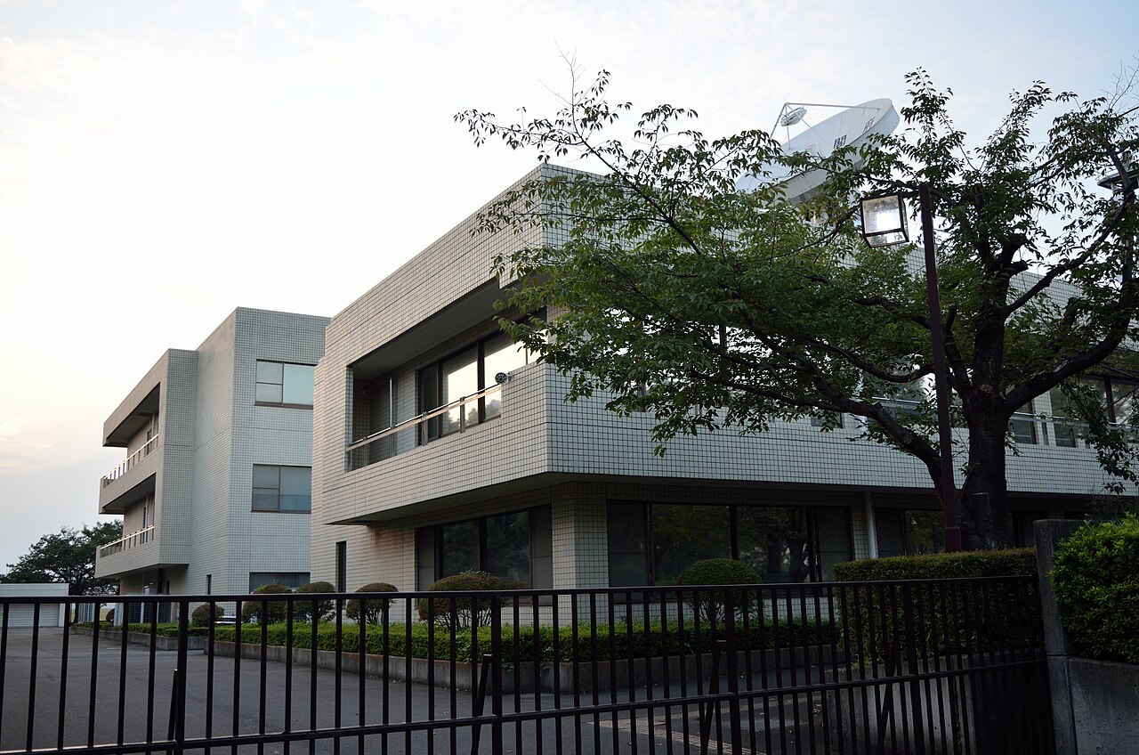 The white building of the national disaster headquarters' reserve facility, seen through trees from outside the gate (2016)