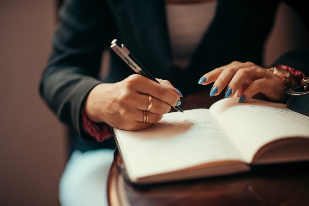 Businesswoman writing in a notebook closeup | オンザショア立川店 立川駅徒歩1分年中無休の溶岩ホットヨガスタジオ ノートにペンで書き込む女性の手元を近くから捉えた横長の写真 立川駅徒歩1分年中無休の溶岩ホットヨガスタジオ